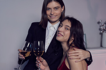 Cropped close-up shot of a two lesbian girls with glasses of white wine. A girl in a black suit hugging her girlfriend in a red dress sitting on a chair in a room. LGBT concept.