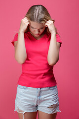 One Beautiful Sad Depressed Caucasian Girl in Pink T-Shirt Suffering From Headache Posing With Both Hands Thinking Against Coral Pink Background.