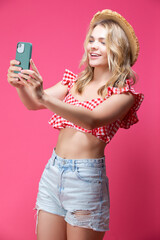 Summer Time Concepts. Smiling and Happy Caucasian Blond Girl in Casual Blue Shirt And Stylish Straw Hat Posing With Smartphone  While taking Selfie Against Coral Background.