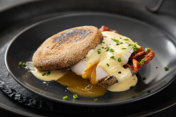 Eggs Benedict mit pochierten Eiern, Sauce Hollandaise, Bacon, Schnittlauch, English Muffin und Cheddar Käse zum Frühstück oder Brunch auf Teller schwarz, Metall Tablet und dunkel Schiefer Hintergrund