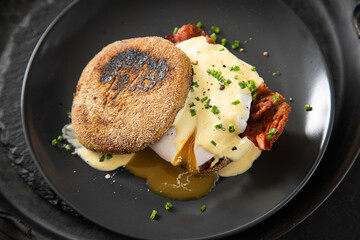 Eggs Benedict mit pochierten Eiern, Sauce Hollandaise, Bacon, Schnittlauch, English Muffin und Cheddar Käse zum Frühstück oder Brunch auf Teller schwarz, Metall Tablet und dunkel Schiefer Hintergrund