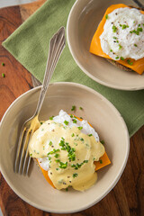 Eggs Benedict vegetarisch mit pochierten Eiern, Sauce Hollandaise, Schnittlauch, English Muffin und Cheddar Käse zum Frühstück oder Brunch auf Teller hell mit Serviette grün, Silber Gabel, Holzbrett u