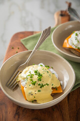 Eggs Benedict vegetarisch mit pochierten Eiern, Sauce Hollandaise, Schnittlauch, English Muffin und Cheddar Käse zum Frühstück oder Brunch auf Teller hell mit Serviette grün, Silber Gabel, Holzbrett u