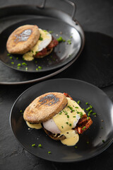Eggs Benedict mit pochierten Eiern, Sauce Hollandaise, Bacon, Schnittlauch, English Muffin und Cheddar Käse zum Frühstück oder Brunch auf Teller schwarz, Metall Tablet und dunkel Schiefer Hintergrund