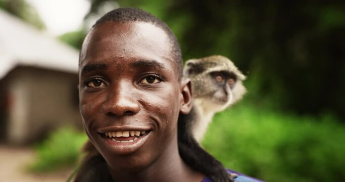 Close Up Portrait of an Authentic African Man Carrying a Little Monkey Looking at the Camera and Smiling with Greenery in the Background. Black Friendly Male With Big Happy Smile. Documentary Footage