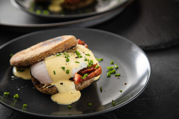 Eggs Benedict mit pochierten Eiern, Sauce Hollandaise, Bacon, Schnittlauch, English Muffin und Cheddar Käse zum Frühstück oder Brunch auf Teller schwarz, Metall Tablet und dunkel Schiefer Hintergrund