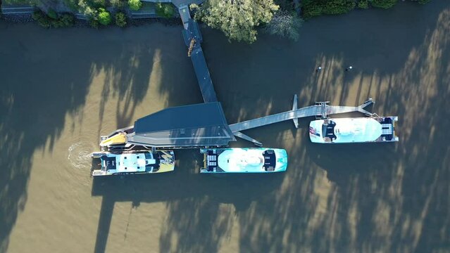 Top down drone shot of City Cat Boat leaving dock and driving away on river. Shot in evening sun with long shadows. 2 other boats are also docked. Shot near UQ, St Lucia, Qld Austalia