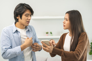 Infidelity, suspicion asian young couple love fight relationship, wife holding cellphone, smartphone cheating on phone, scolding husband about mistrust, distrust and jealousy when sitting at home.