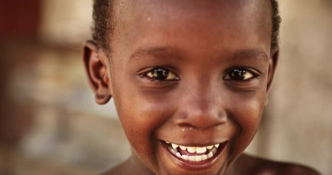 Close Up Portrait of a Shy Authentic African Boy Smiling and Laughing at the Camera with a Blurred Background. Black Male Kid Representing Future, Hope, and Acceptance. Documentary Footage