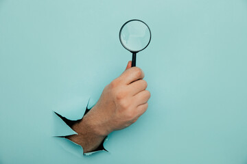 Male hand holds black magnifying glass in torn hole of blue background. Searching information concept