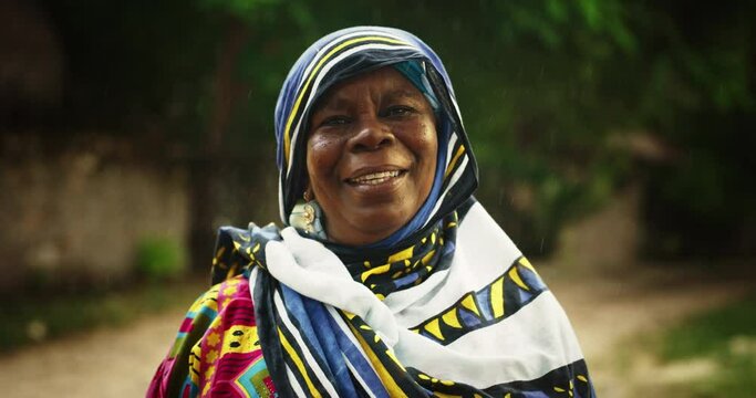 Portrait of Authentic Old African Woman Standing Under the Rain, Looking at the Camera and Smiling with a Blurry Greenery Background. Female in Traditional Clothes Enjoying the Blessing of Rain