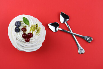 Anna Pavlova dessert with berries and a leaf of mint on red background with heart shaped spoons nearby. Saint Valentines day food. Romantic decoration of the table. White protein cupcake for a couple