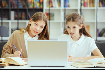 School children wearing uniform sitting at desk, looking at laptop and writing in exercise books,...