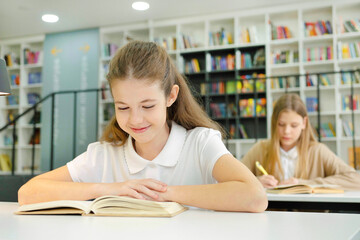 Happy schoolgirl sitting in class and reading book, student behind her writing in notebook, bookshelves on background. Children studying in school. Concept of education