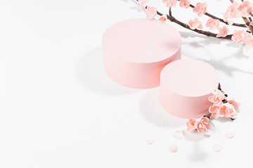 Spring festive stage mockup with two pink cylinder podiums for presentation cosmetic products, goods, and branch of tender pink sakura flowers on white table in traditional japanese style, top view.