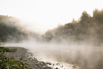 Idyllic gentle morning summer landscape with fog on river with white  haze flow above water, silhouette green forest on slopes in bright golden sunlight. Amazing misty wild nature, tourism outdoors.
