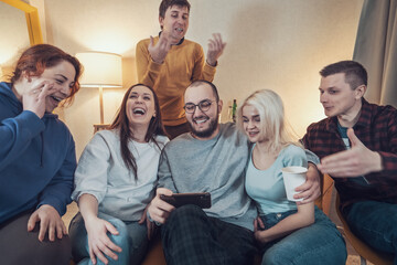 Group of cheerful young friends looking at smartphone at home party, watching online video or making selfie or video call.