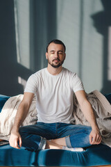 Pleased young man with closed eyes sitting on sofa in lotus pose meditating at home dressed in casual clothes. People meditation wellness concept. - obrazy, fototapety, plakaty