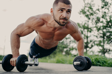 Fototapeta na wymiar Horizontal shot of muscular unshaven man does push up exercises, leans at barbells, focused into distance, puts all efforts while trains abdominal muscles poses outdoor. Sporty lifestyle concept