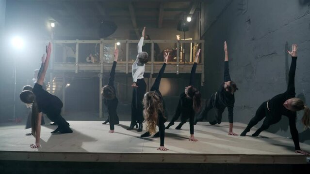 Young fit actors and actresses in plank position on backstage raising hands one by one looking up. Beautiful stage director talking as men and women rehearsing play in theater indoors