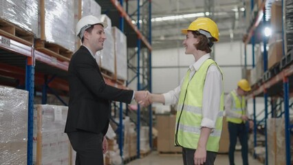 businessman shaking hands with worker wearing hardhat standing against warehouse shelves in background.worker wearing reflective jacket holding QR code scanner standing in factory warehouse smiling.