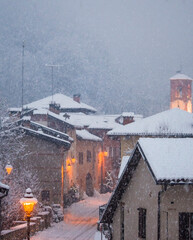 Beautiful evening snowfall in a small typically Italian rural village with the bell tower and houses lit by elegant wall lamps, Italy