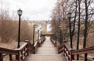 Fototapeta na wymiar Wooden high staircase for descent from the mountain to the city. Lanterns, massive railings. Old trees and shrubs. Below you can see the streets and houses of the townspeople. Early spring.