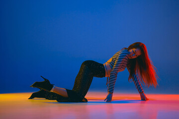 Self-expression. Young girl dancing high heel dance in stylish clothes over blue background in neon...