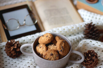 Cup of tea, books, tablet, bowl of cookies, various spices, pine cones and lit candles. Hygge at home. Selective focus.
