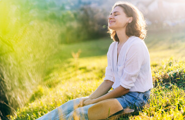 Happy pretty young cheerful woman in white shirt sitting on grass enjoying sunny spring day with eyes closed - 562074269