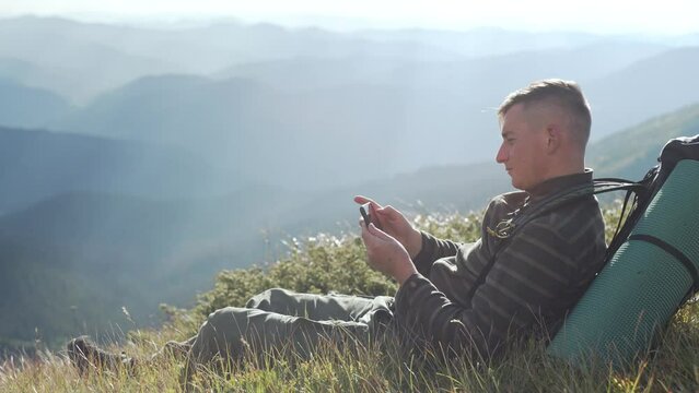 A man with a backpack takes a picture of the landscape on his phone while hiking. Portrait of a guy taking a photo with a smartphone in nature. Sunny day at the top. High quality 4k footage