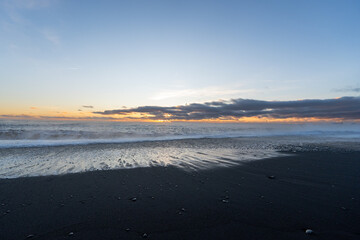 Reynisfjara black sand beach illuminated by the last rays of sunset sun from the horizon of the sea illuminating the clouds