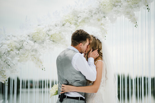The bride and groom kissing. Newlyweds with a bouquet standing on wedding ceremony under the arch decorated with flowers and greenery of the outdoor.