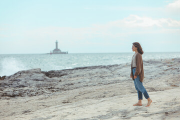 woman walking by rocky sea beach in wet jeans lighthouse on background