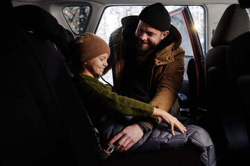 Happy dad putting his daughter in the back seat of car and fastening her with seat belt before trip