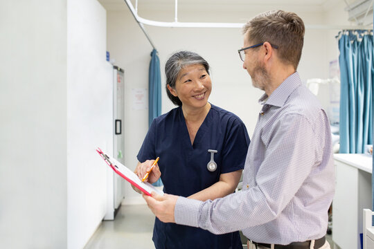 smiling middle aged woman wearing blue scrubs holding a file while talking to a man in a clinic ward