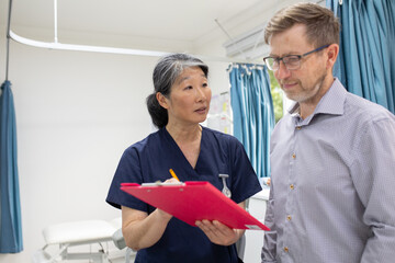 middle aged woman wearing blue scrubs holding a file while talking to a man in a clinic ward