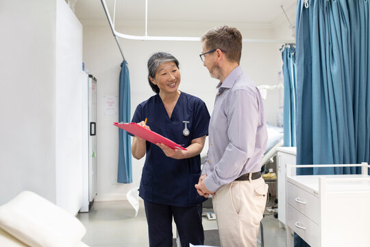 smiling middle aged woman wearing blue scrubs holding a file while talking to a man in a clinic ward