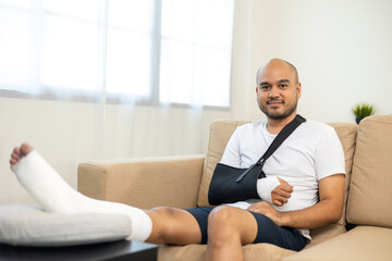 Happy man recovery from accident fracture broken bone injury with leg splints in cast neck splints collar arm splints sling support arm in living room. Social security and health insurance concept.