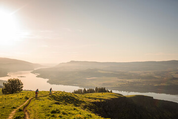 COLUMBIA RIVER GORGE, OR, USA. Two young women stop to rest on mountain bikes while riding a single-track trail through open meadow with river and volcano in distance.