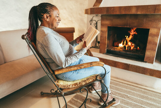 Senior african woman reading book in front of cozy fireplace at home - Winter lifestyle concept - Focus on face