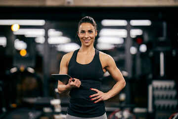 A fit muscular female personal trainer is holding tablet in her hands and smiling at the camera in a gym. - Powered by Adobe