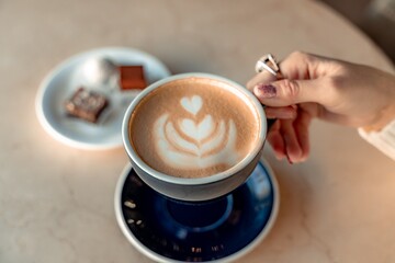 A cup of fresh cappuccino coffee in the hands of a woman on a fashionable background of a white marble table, next to a plate with sweets. Coffee addiction. Top view, flat lay.