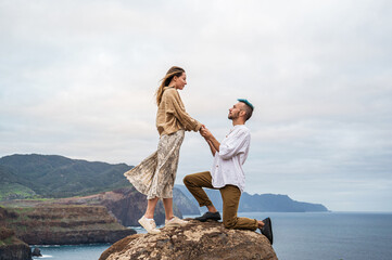 A young couple stands on a rocky cliff by the ocean, with the man down on one knee proposing to the woman