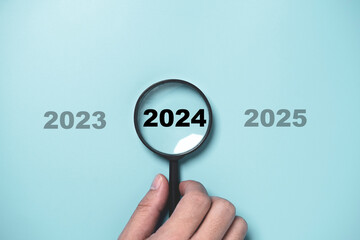 Hand holding black magnifier glass with 2024 year among 2023 and 2025 for preparation and focus ...