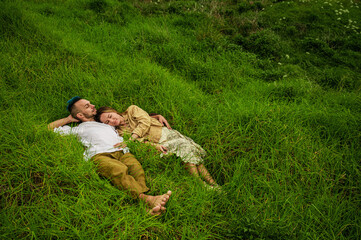 A young couple lies in a field of lush green grass, embracing each other in a hug.