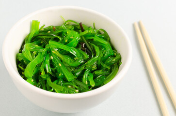Hiyashi Wakame Chuka or seaweed salad in a white bowl with bamboo sticks on a gray background. The concept of traditional oriental food. Vegetarian food. Horizontal orientation. Selective focus.