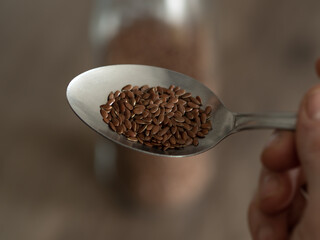 Close-up of a spoon filled with flax seed, to eat some seeds, large quantity of flax seed in a glass container. Linseed reserve on wood table.