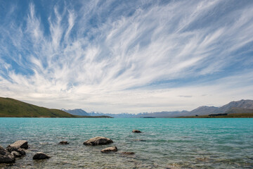 Wispy clouds and blue skies over the turquoise blue water of Lake Tekapo in New Zealand with snow capped mountains in the distance and rocks in the foreground