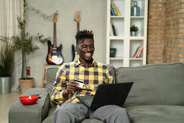 Cheerful young man paying bills online with credit card and laptop. African man using credit card...